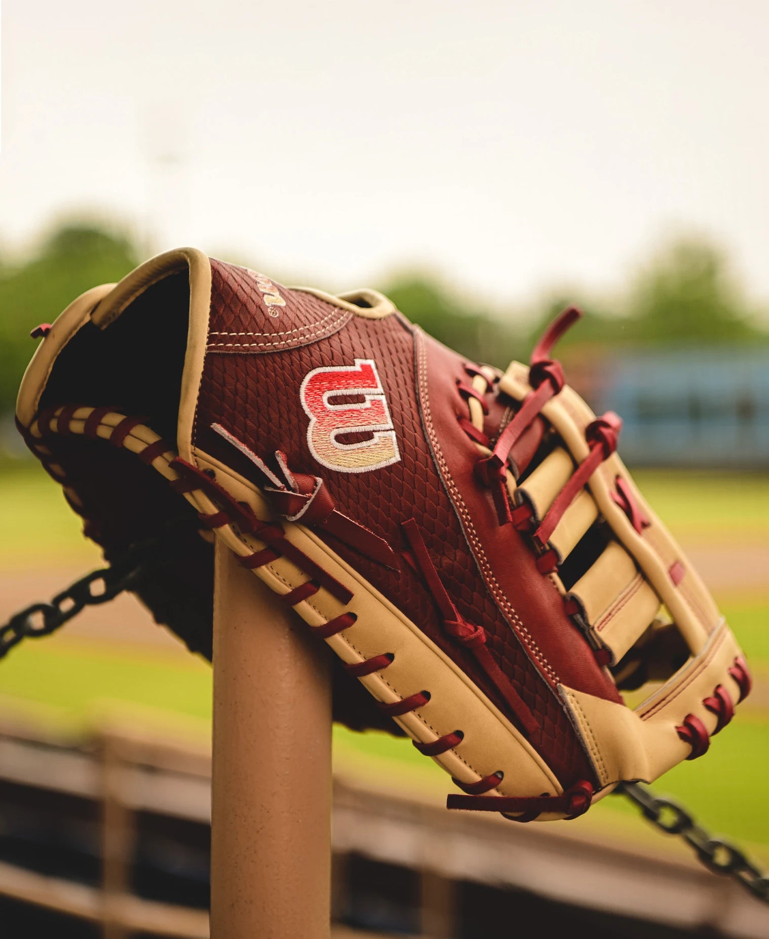 A Wilson A2K 1679 12.5" Baseball First Base Mitt (WBW102684125), brown and tan with a red and white W logo and Single Post Web, hangs on a metal post against a blurred field and greenery background.