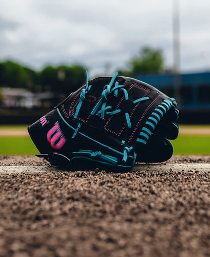 The Wilson A2K B2 12" Baseball Glove - SPRING 2025 (WBW10269212) features Pro Stock Select leather, a Closed 2-Piece Web, black color with blue laces and pink accents, and rests on the mound with trees and a blue fence in the background.