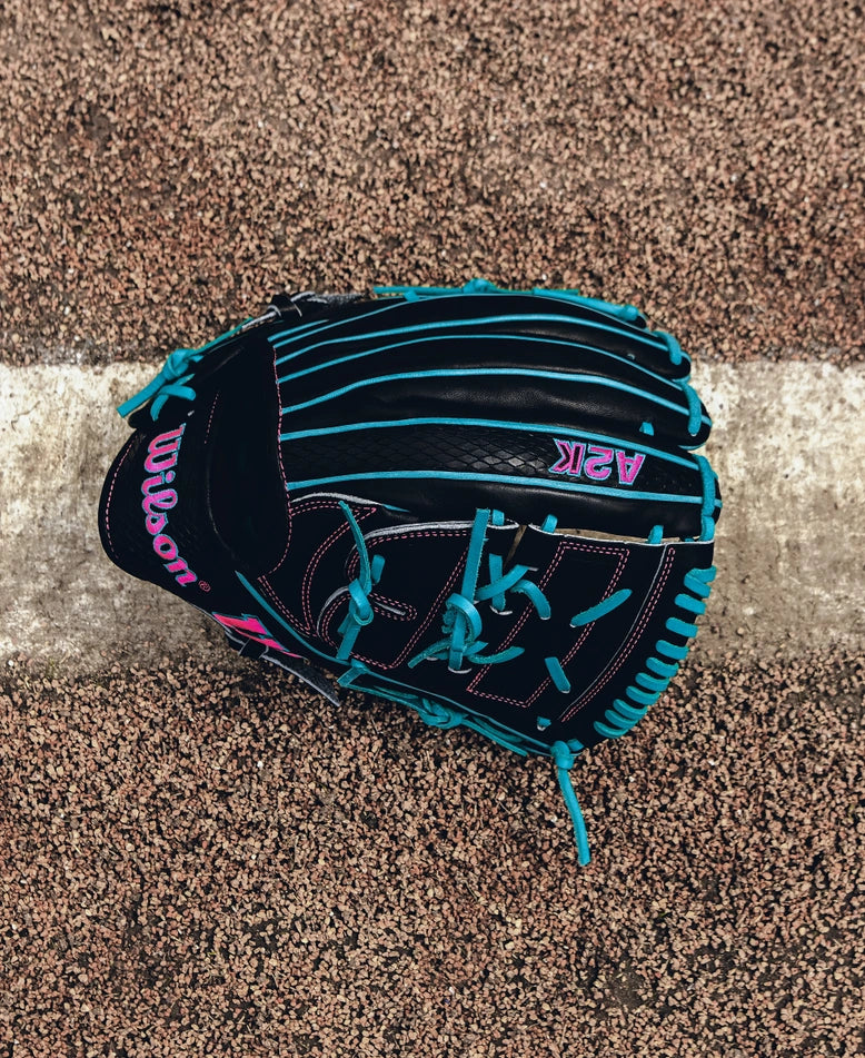 A Wilson A2K B2 12" Baseball Glove (SPRING 2025: WBW10269212) in black Pro Stock Select leather with turquoise laces and pink lettering rests on a brown and white textured surface over a white line.