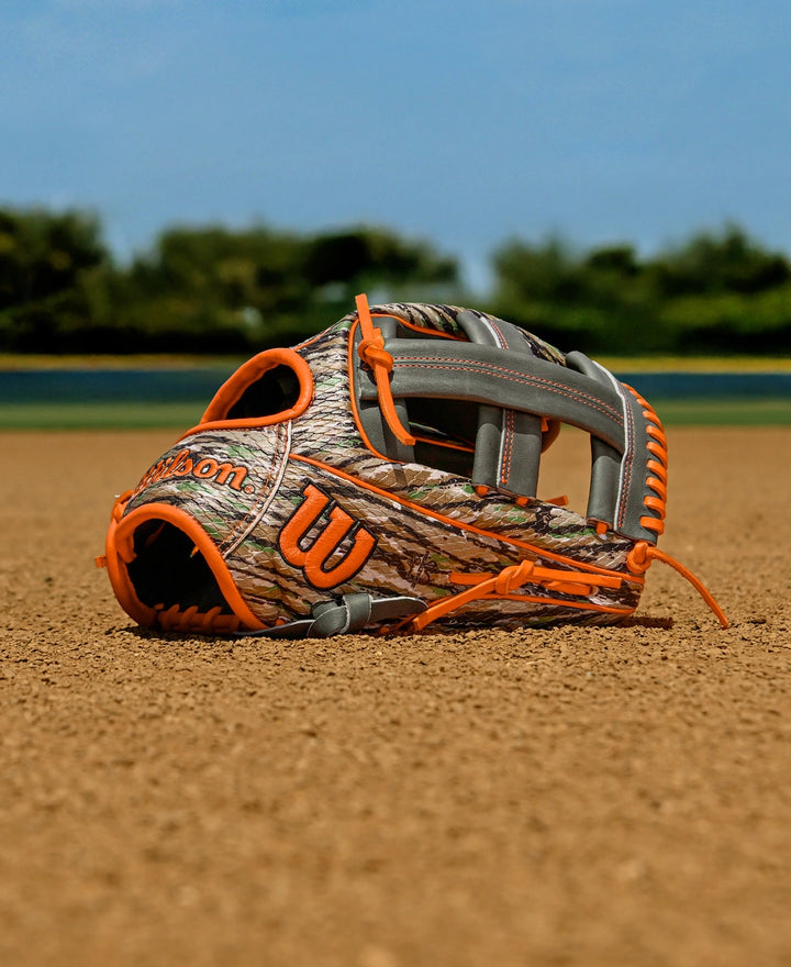 A Wilson A2000 GH2 11.75" Gunnar Henderson GM Baseball Glove (WBW1034511175) with a camouflage pattern, orange and gray accents, rests on the infield dirt, framed by green trees and a blue sky.