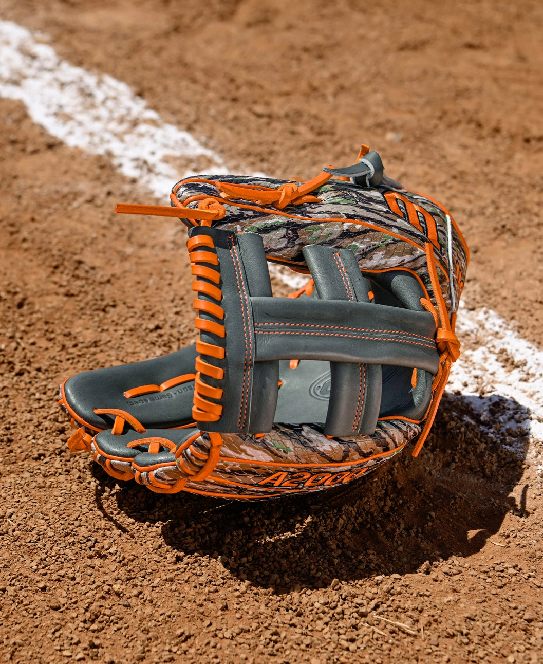 A Wilson A2000 GH2 11.75" Gunnar Henderson GM Baseball Glove (WBW1034511175) with orange and camouflage details lies open on a dirt field near a white chalk baseline.