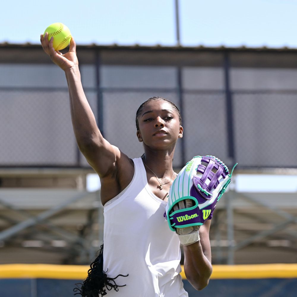 A young woman in a white tank top throws a yellow softball while wearing the Wilson A2000 FP50SS 12.25" Fastpitch Glove (FALL 2025: WBW1034891225) outdoors on a sunny day, with a fence and bleachers behind her.