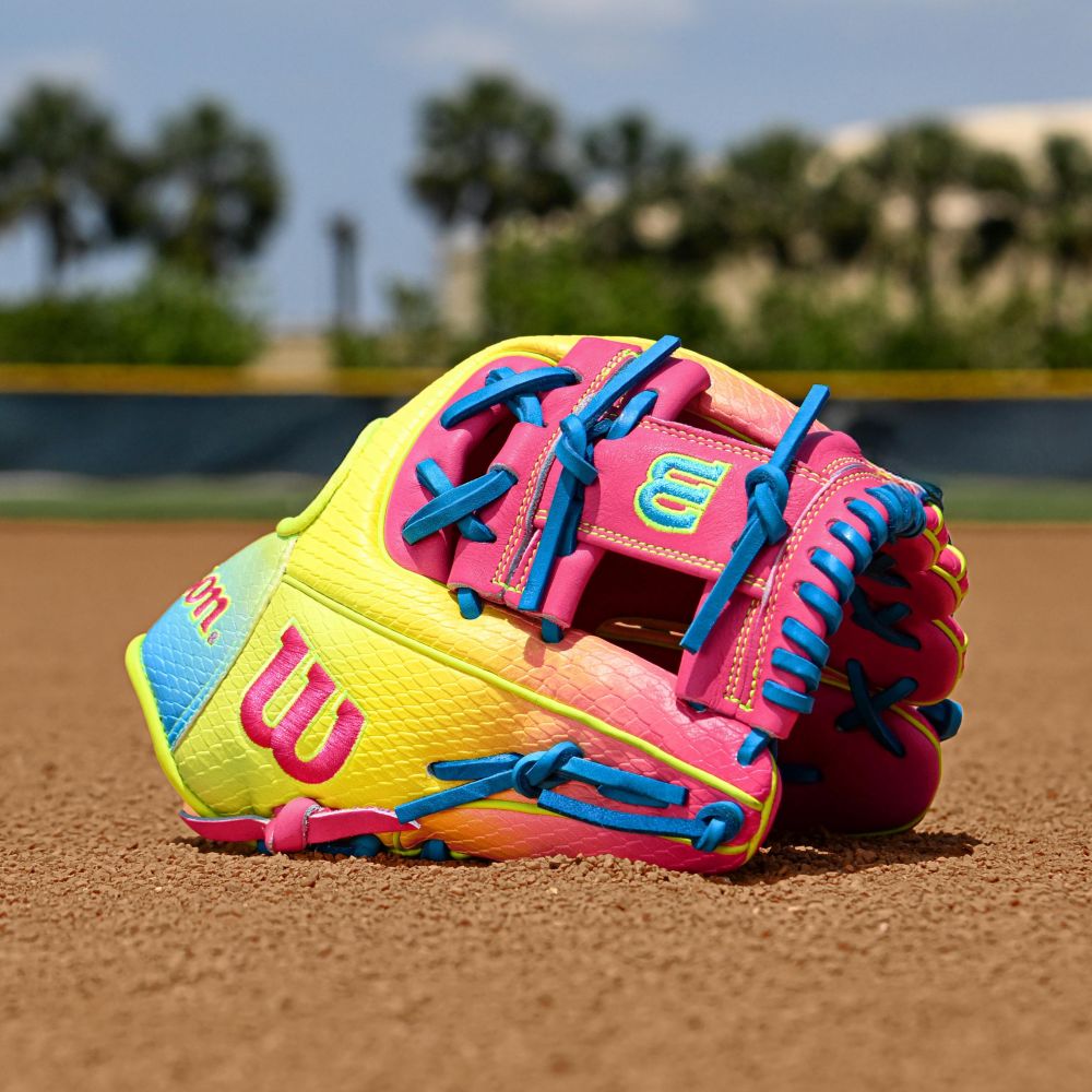 A Wilson A2000 1786SS 11.5" Baseball Glove (WBW103492115) with yellow, pink, and blue accents lies on a dirt field, trees and a blurred building in the background.