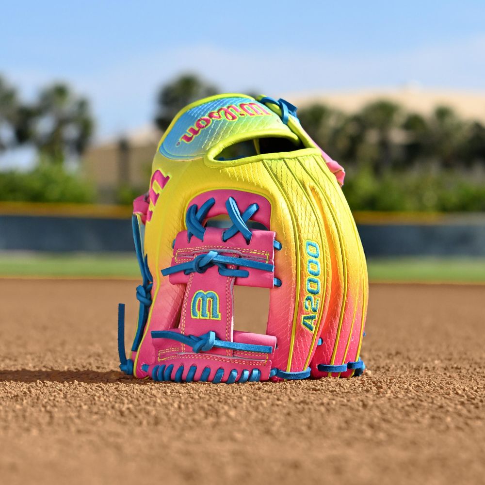 The Wilson A2000 1786SS 11.5" Baseball Glove (WBW103492115) features bright pink, yellow, and blue accents and is shown upright on a dirt field, with a blurred outfield and trees behind it.