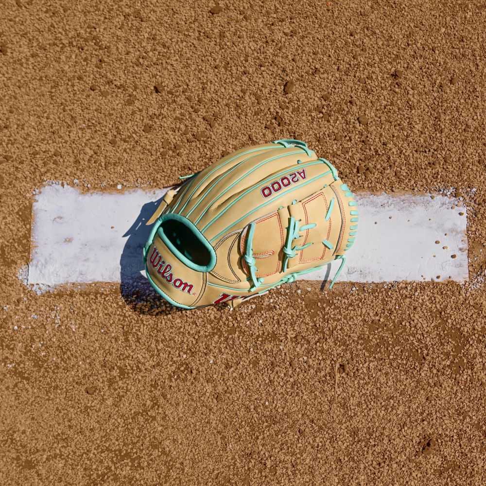A Wilson A2000 B12 12" Baseball Glove in tan and mint green Pro Stock leather (WBW10349812) rests on a white chalk baseline of a dirt baseball field.