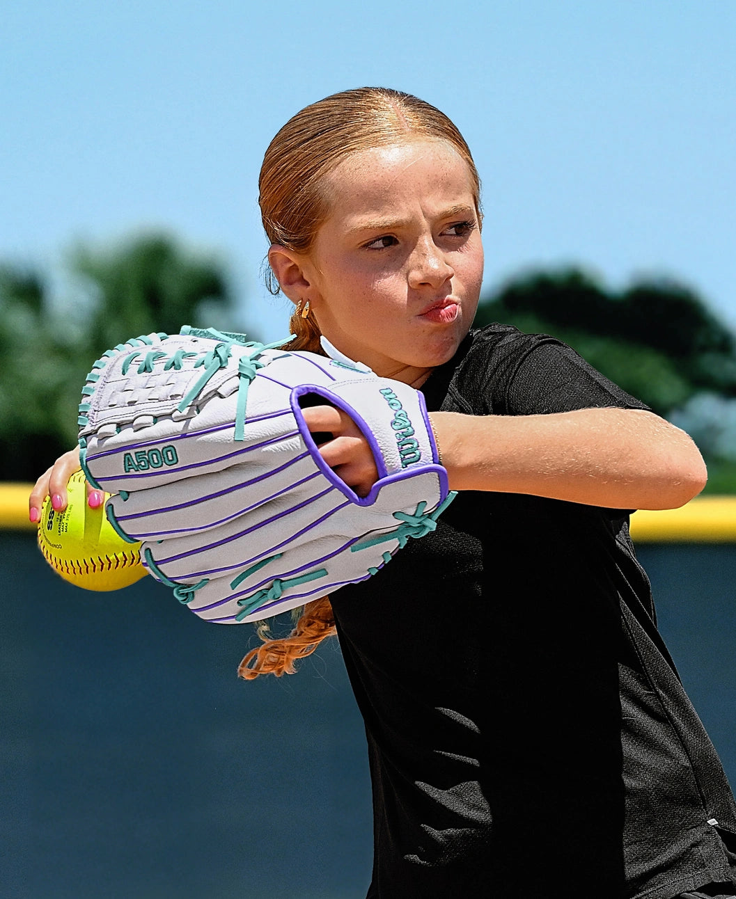 A young girl with light brown hair in a ponytail prepares to throw a softball outdoors on a sunny day, wearing a black shirt and the Wilson A500 Siren 11.75" Fastpitch Glove featuring teal details and Quick-Fit Wrist.