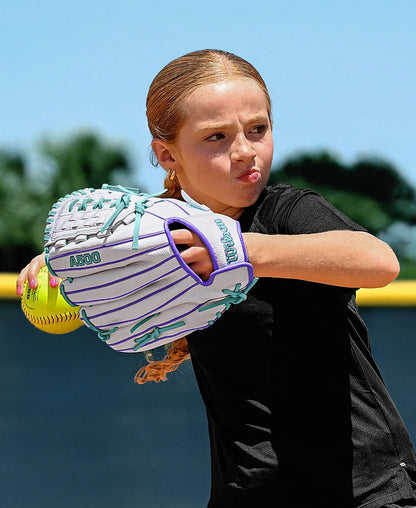 A young girl with light brown hair in a ponytail prepares to throw a softball outdoors on a sunny day, wearing a black shirt and the Wilson A500 Siren 11.75" Fastpitch Glove featuring teal details and Quick-Fit Wrist.