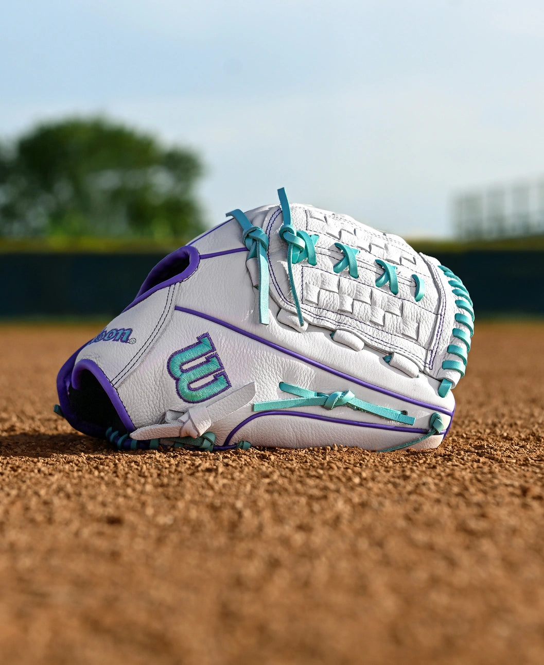 A Wilson A500 Siren 11.75" Fastpitch Glove (WBW1035581175) in white and teal top grain leather lies on the brown infield dirt of a baseball field, with green grass, trees, and an outfield fence softly blurred in the background.
