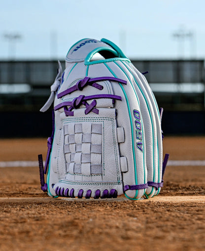 The Wilson A500 Siren 12" Fastpitch Glove (WBW10355912) is a white youth glove with teal and purple accents, crafted from top grain leather, standing upright on a dirt field with a blurred stadium fence and lights behind it.