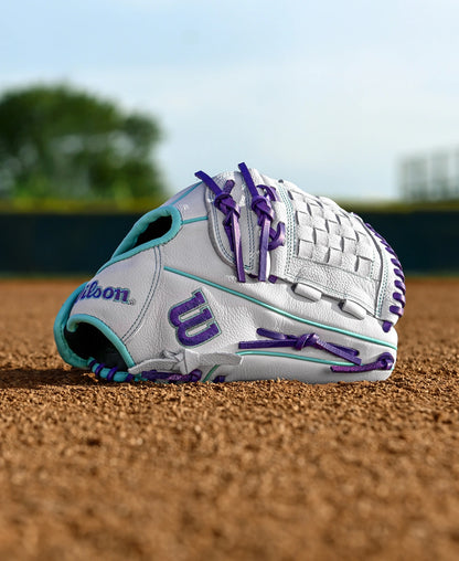 A Wilson A500 Siren 12" Fastpitch Glove (WBW10355912) in white with purple and teal accents and a quick-fit wrist rests on the dirt of a baseball field, with green trees and a blurred background behind it.