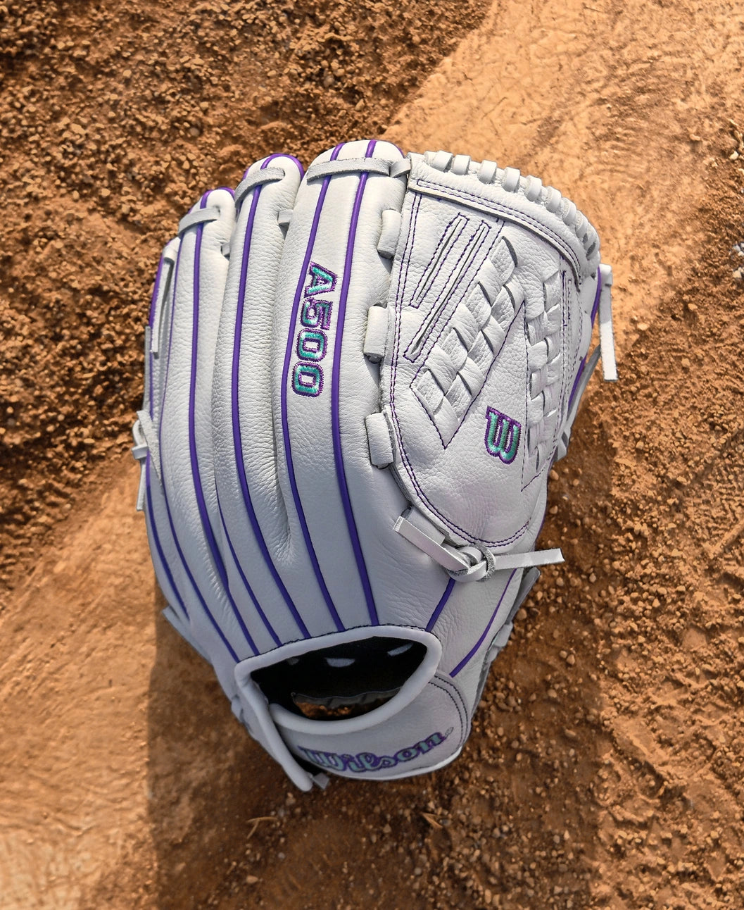 A Wilson A500 Siren 12.5" Fastpitch Glove (WBW103561125) with purple accents and a Quick-Fit Wrist rests on a dirt infield.