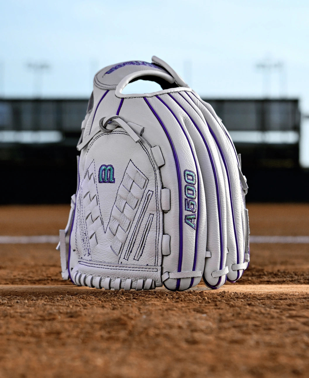 The Wilson A500 Siren 12.5" Fastpitch Glove (WBW103561125), a white top grain leather youth glove with purple stitching and A500 embroidery, stands upright on a dirt infield against a blurred stadium and sky backdrop.