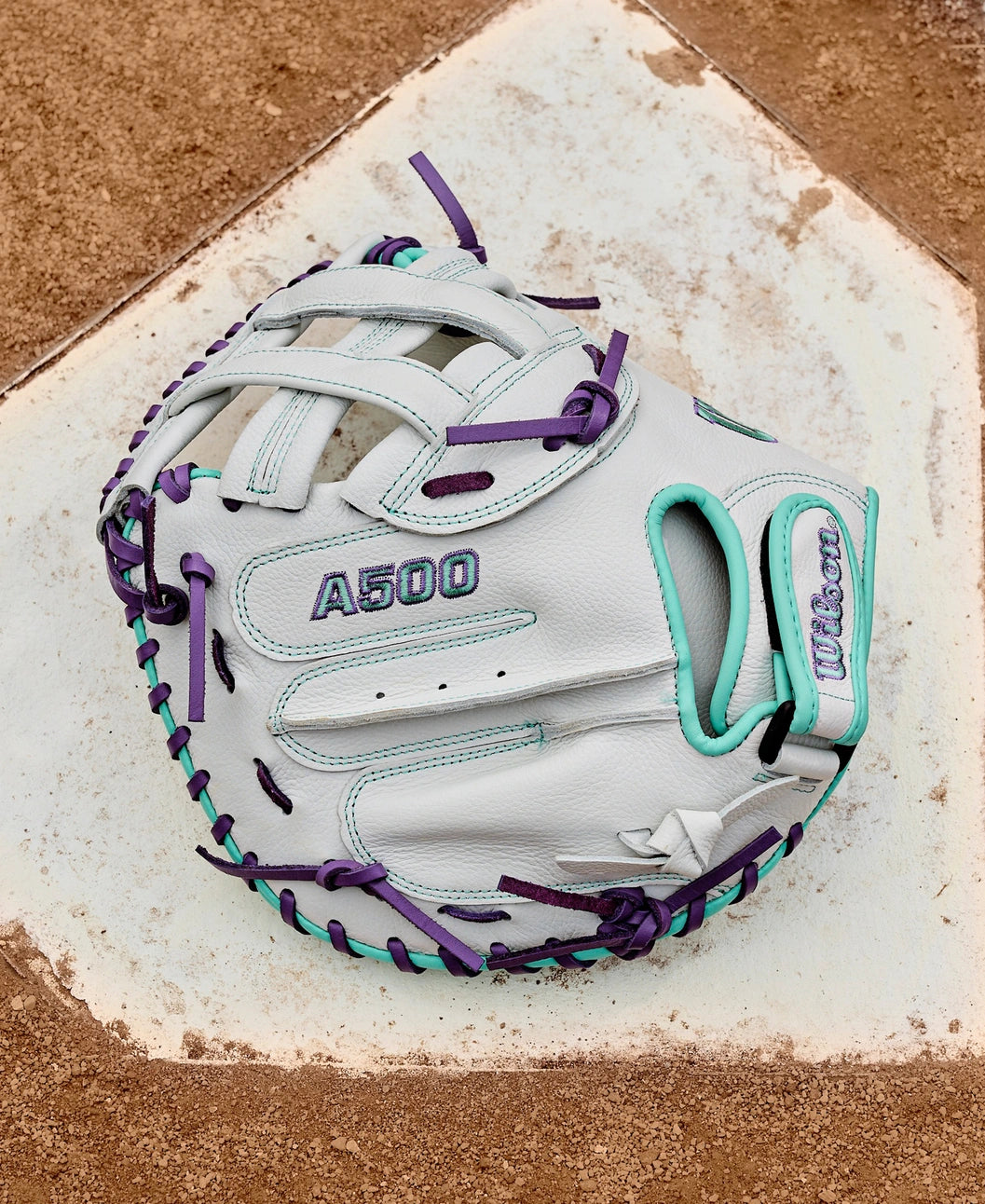A Wilson A500 Siren 32" Fastpitch Catcher's Mitt (WBW10356332) in white with teal and purple laces sits on home plate on a dirt baseball field.