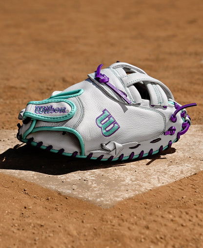 A Wilson A500 Siren 32" Fastpitch Catcher's Mitt (WBW10356332), white with teal and purple accents, rests on home plate of a dirt field.
