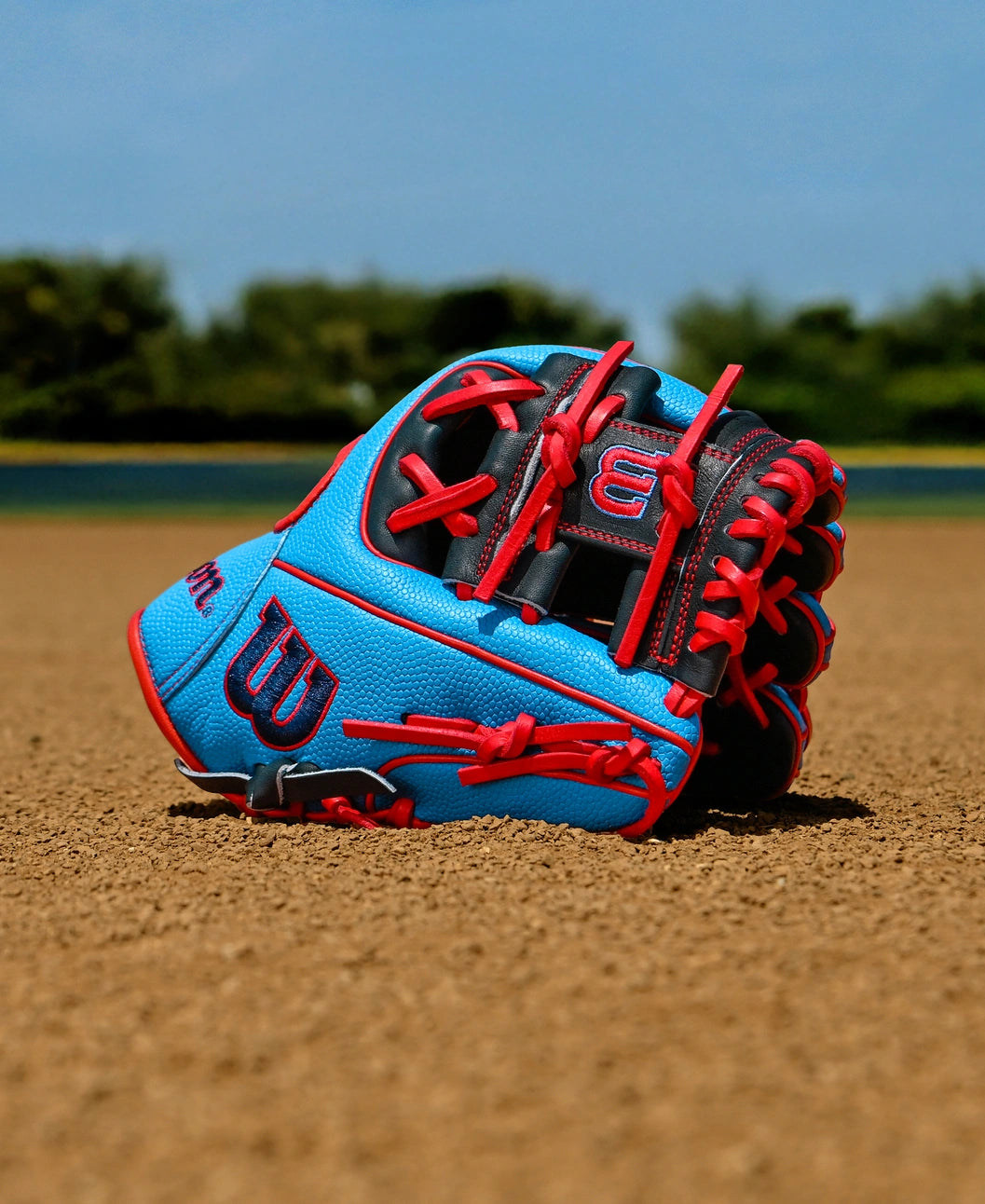 A Wilson A2000 PF11SS 11" Baseball Glove (WBW10394011) with red laces lies on a dirt infield, backed by green trees and a clear blue sky.