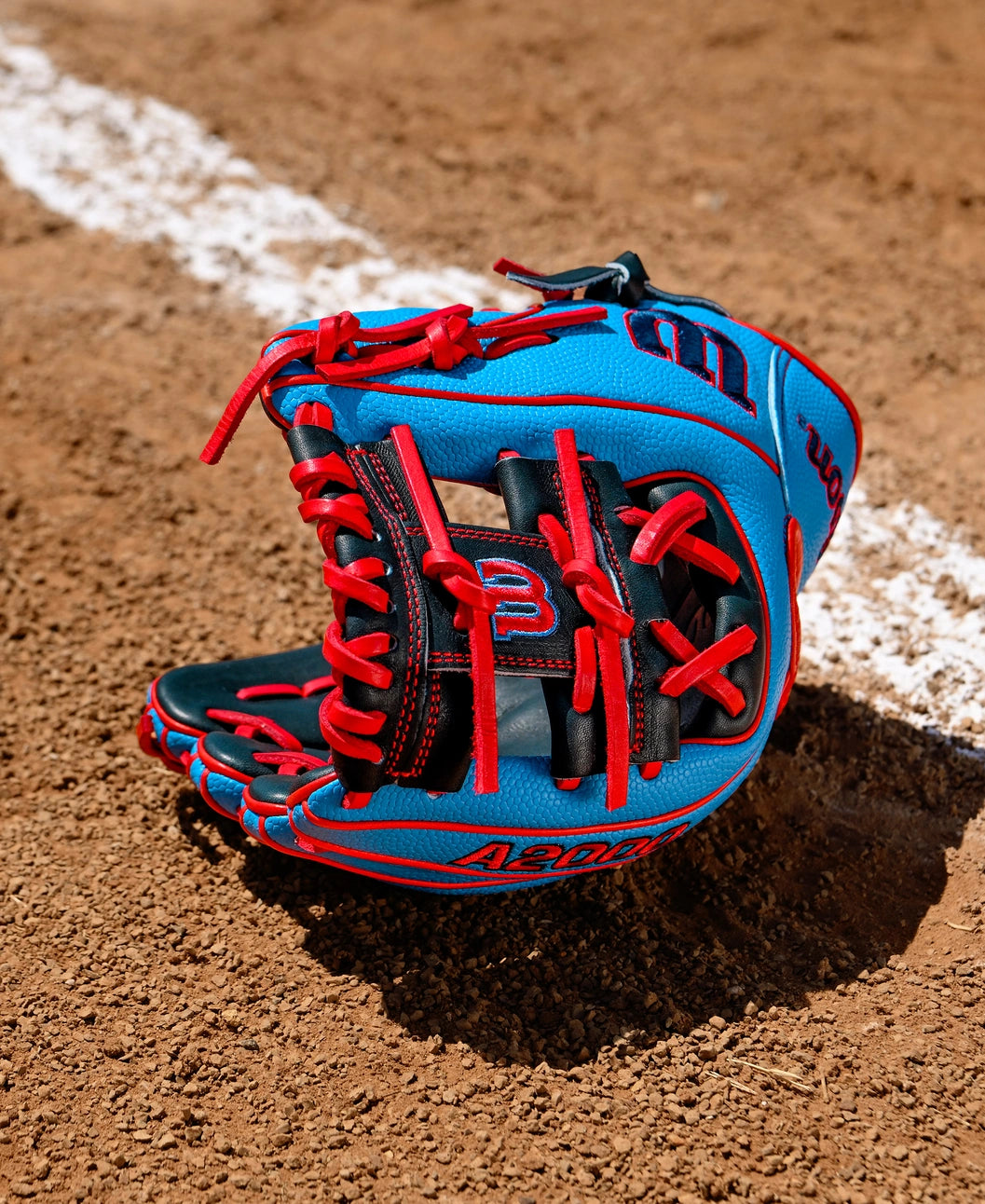 A Wilson A2000 PF11SS 11" Baseball Glove by Wilson, featuring blue and black colors with red laces and Pro Stock leather, rests on a dirt field near a white chalk baseline.