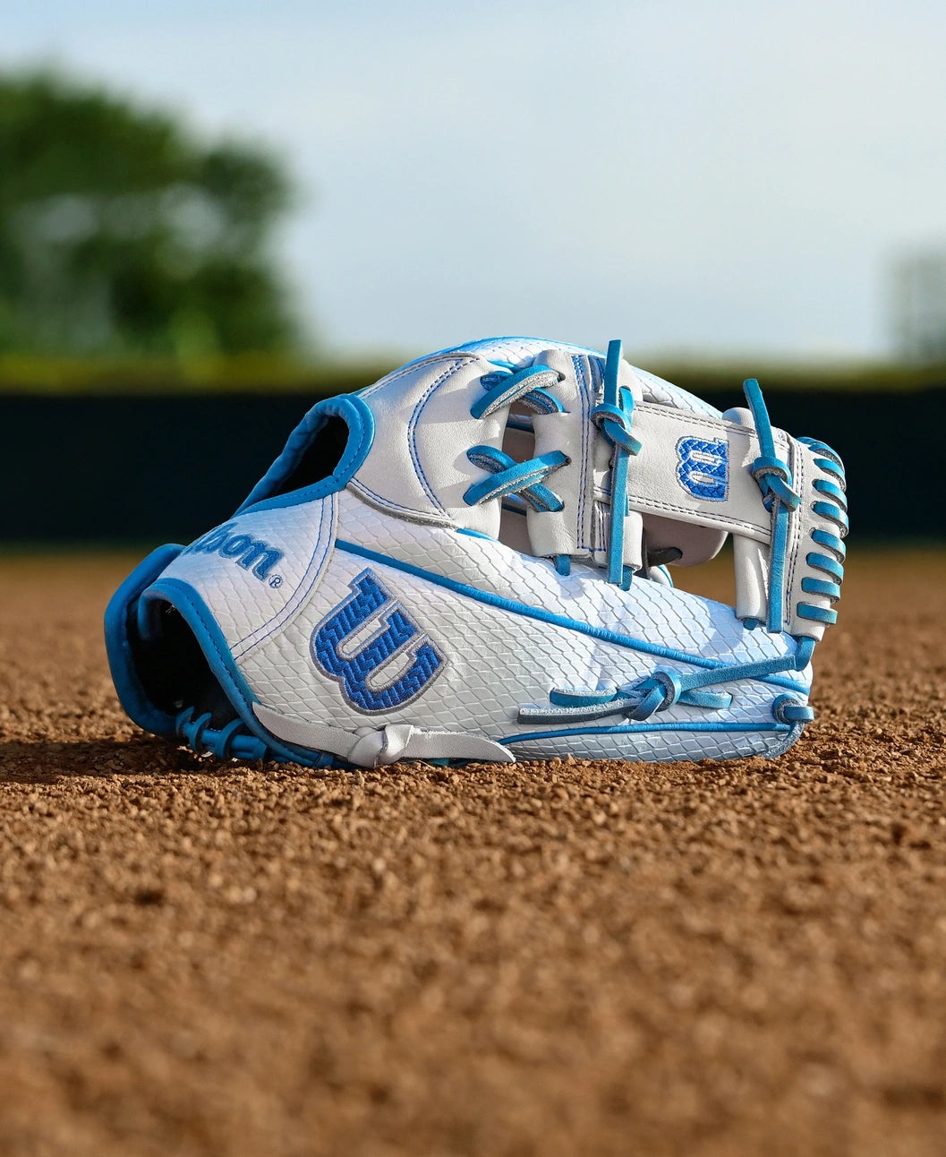 The Wilson A2000 H12SS 12" Fastpitch Glove (WBW10394612) by Wilson, white and blue, rests on a baseball field’s dirt with green grass and trees softly blurred in the background.
