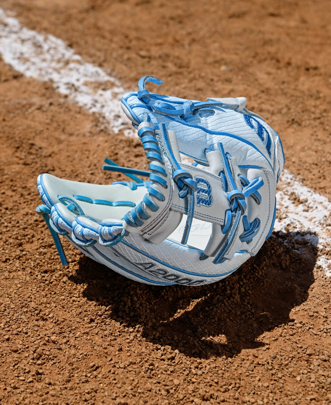 A Wilson A2000 H12SS 12" Fastpitch Glove (WBW10394612) in light blue and white, featuring Pro Stock leather and blue laces, rests open on reddish-brown dirt near a white foul line on a baseball field.