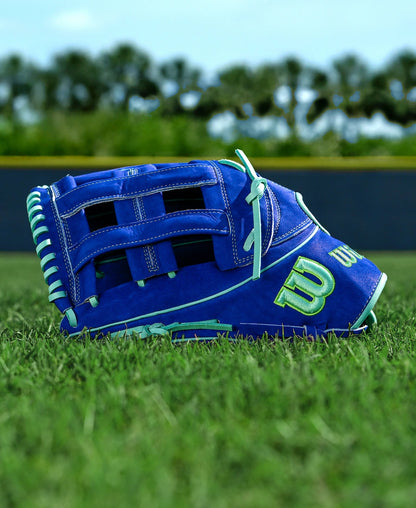 A Wilson A2K JS22 12.75" Juan Soto GM baseball glove, featuring Pro Stock Select leather with blue, white, and green accents, rests on green grass. A fence and trees blur in the background beneath a clear sky.