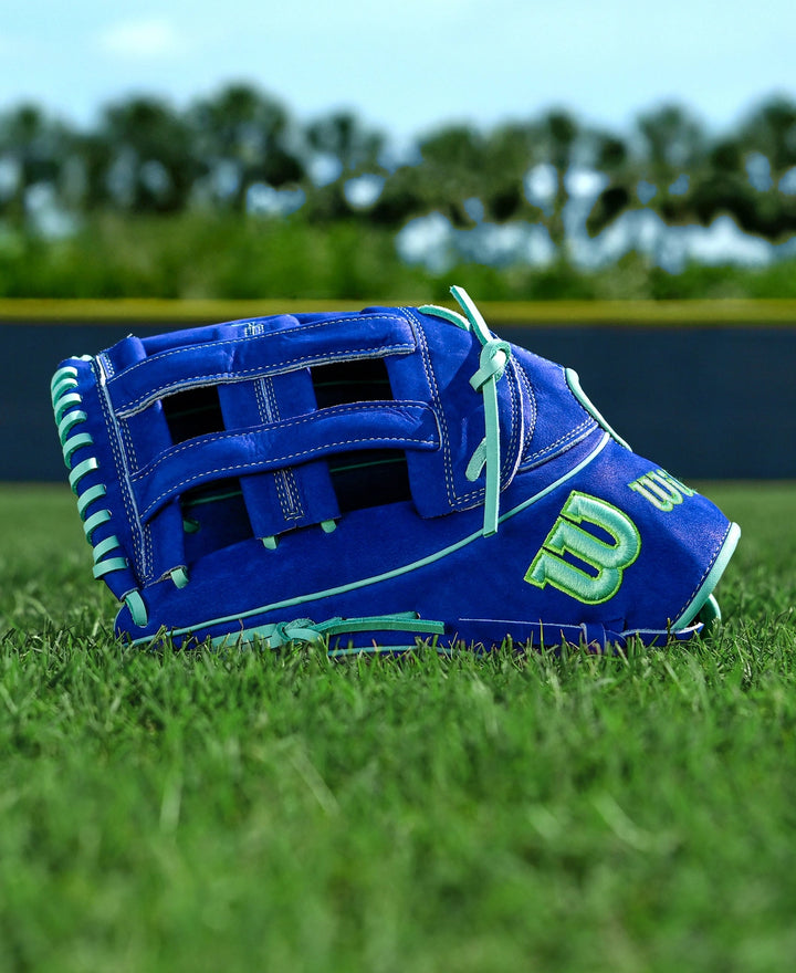 A Wilson A2K JS22 12.75" Juan Soto GM baseball glove, featuring Pro Stock Select leather with blue, white, and green accents, rests on green grass. A fence and trees blur in the background beneath a clear sky.