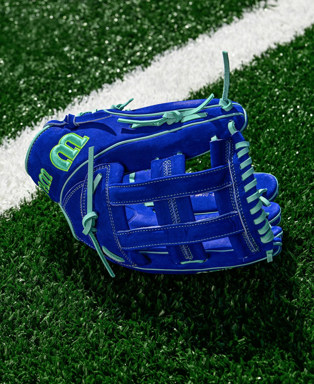 The Wilson A2K JS22 12.75" Juan Soto GM Baseball Glove in blue with green laces and accents, crafted from Pro Stock Select leather, rests on artificial turf beside a white line.
