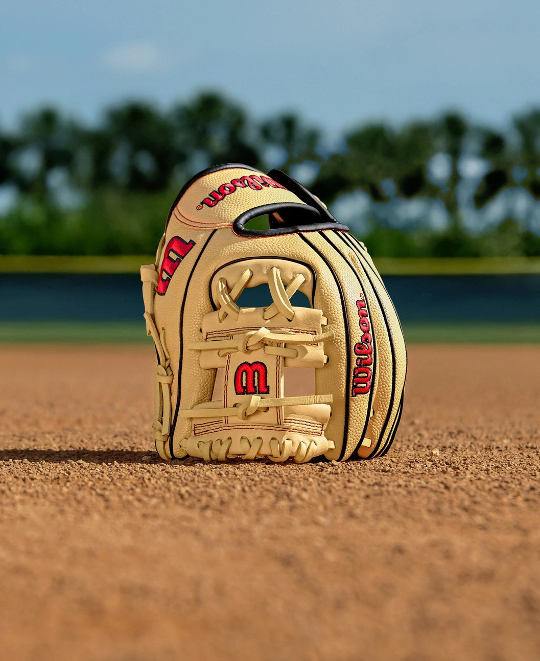 A Wilson A1000 PF11 11" tan infield baseball glove (WBW10395511) with red lettering stands upright on the infield dirt, trees and a blue sky blurred in the background.