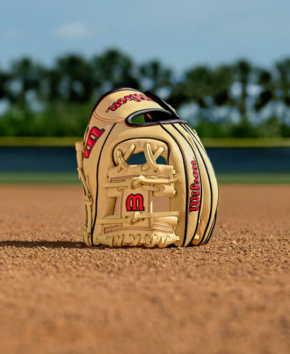 A Wilson A1000 PF11 11" tan infield baseball glove (WBW10395511) with red lettering stands upright on the infield dirt, trees and a blue sky blurred in the background.