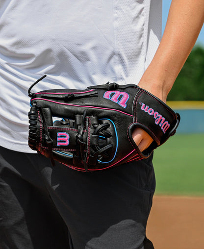 A person in dark pants and a white shirt stands on a baseball field, holding a Wilson A1000 DP15 11.5" Baseball Glove (WBW103956115) by Wilson in their right hand, with the field and greenery visible in the background.