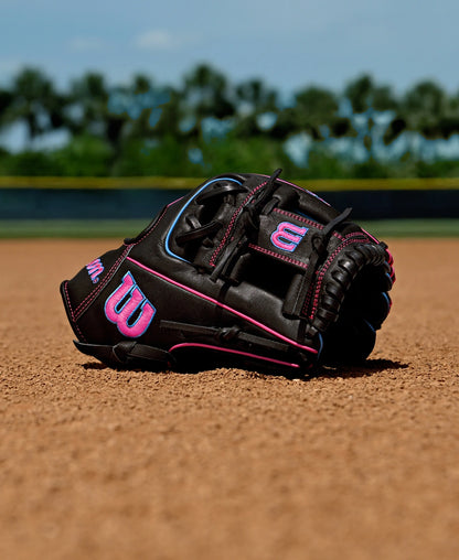 A Wilson A1000 DP15 11.5" Baseball Glove (WBW103956115) by Wilson, featuring black leather with pink and blue accents, rests on the dirt with green grass and blurred trees in the background.
