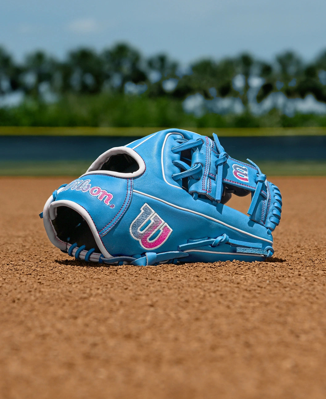 A Wilson A1000 1786 11.5" Baseball Glove (WBW103957115) by Wilson, featuring bright blue with pink and white accents, rests on infield dirt with blurred green trees and a blue sky in the background.