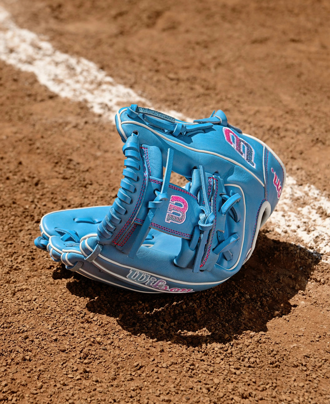 The Wilson A1000 1786 11.5" Baseball Glove (WBW103957115), featuring blue with white and pink accents, lies on the dirt near a chalk baseline. Crafted from durable Pro Stock leather for lasting performance.