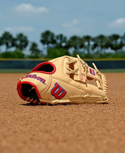 A Wilson A1000 1975 11.75" Baseball Glove (WBW1039591175) by Wilson, tan with red and blue accents, rests on a baseball field’s dirt infield, green trees and blue sky softly blurred in the background.