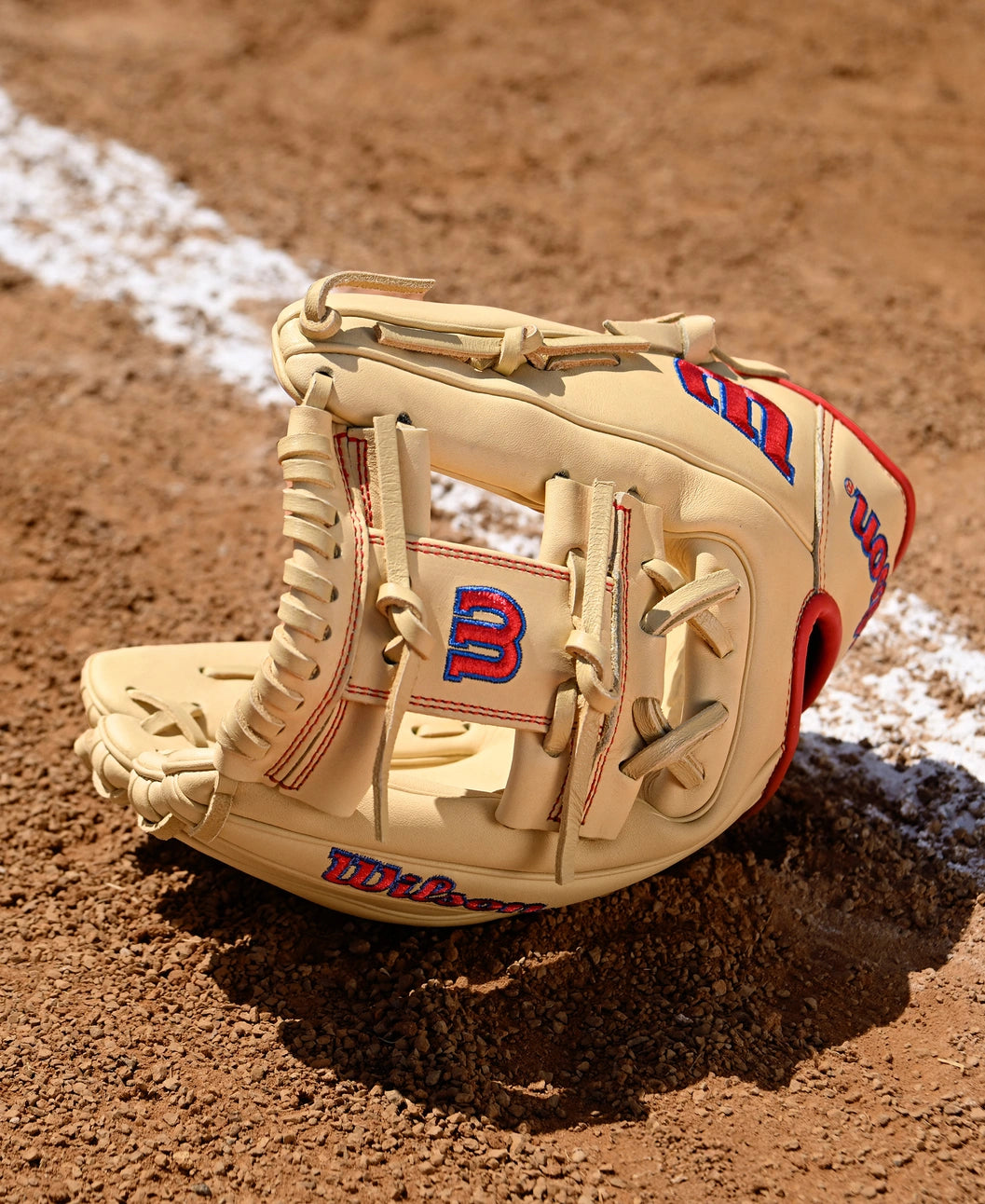 A Wilson A1000 1975 11.75" Baseball Glove (WBW1039591175) by Wilson, featuring tan leather with red and blue stitching, rests open on a dirt field near a white chalk line.