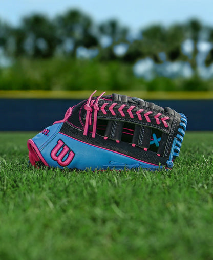 A Wilson A1000 1892 12.25" Baseball Glove: WBW1039621225 with blue, pink, and black accents rests on green grass. The Full Grain leather glove stands out against a blurred backdrop of trees and a blue outfield fence.