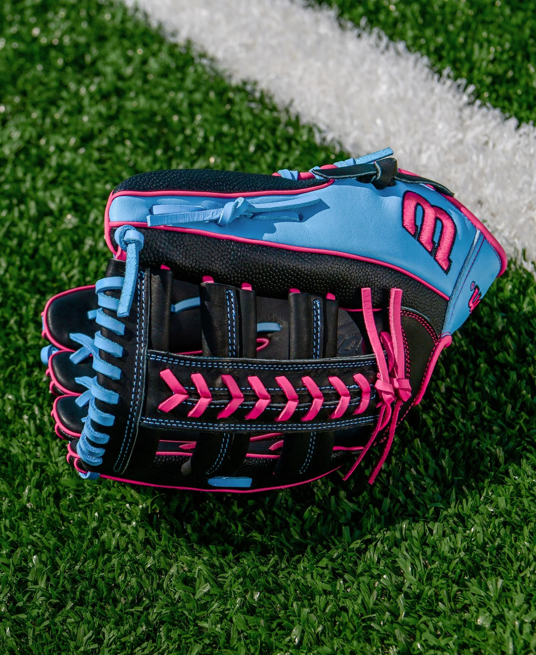 A Wilson A1000 1892 12.25" Baseball Glove (WBW1039621225) in black with pink and light blue accents, made from Full Grain leather, sits on green artificial turf near a white line.