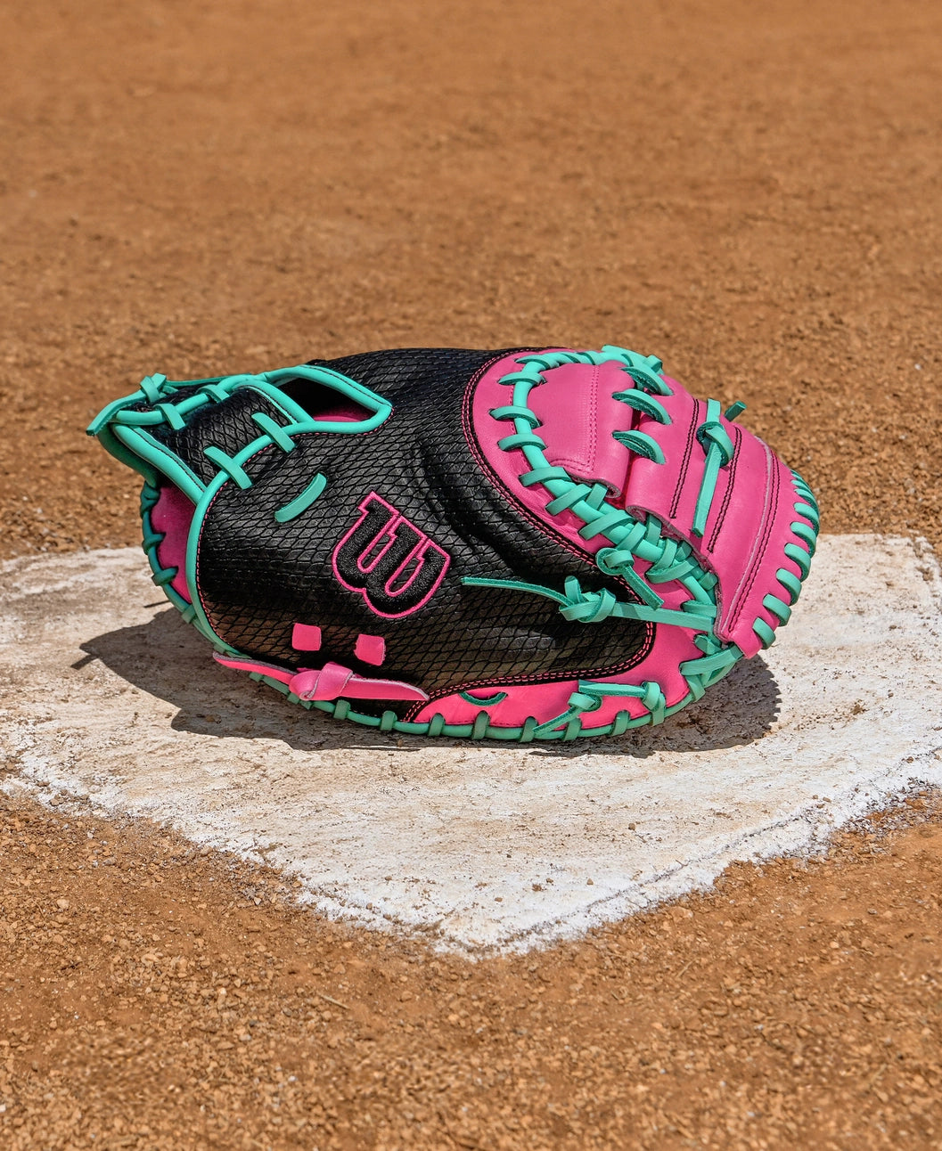 The Wilson A1000 CM33 33" Catcher's Mitt by Wilson, crafted from black full-grain leather with pink and teal laces, rests on home plate, adding a vibrant touch to the dirt baseball field.