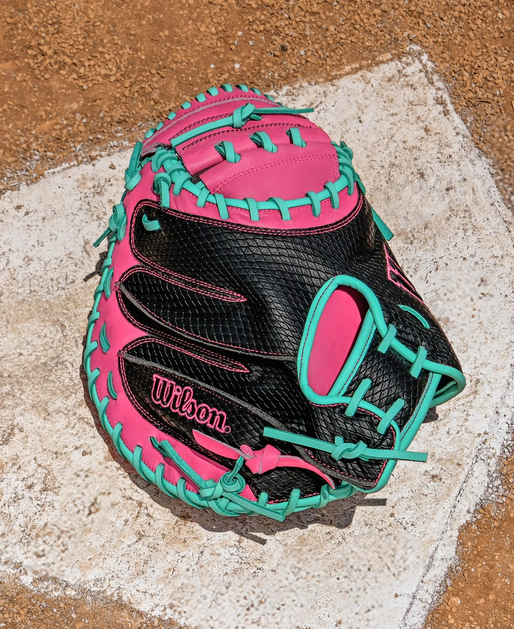 A Wilson A1000 CM33 33" Baseball Catcher's Mitt (WBW10396633) in black, pink, and teal full grain leather rests on a white home plate with a dirt background.