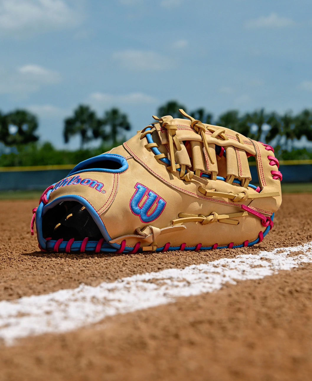 A Wilson A1000 1620 12.5" First Base Mitt by Wilson, in tan and blue full grain leather with pink laces, lies on the dirt near a white foul line on a baseball field, with green trees and a blue sky in the background.