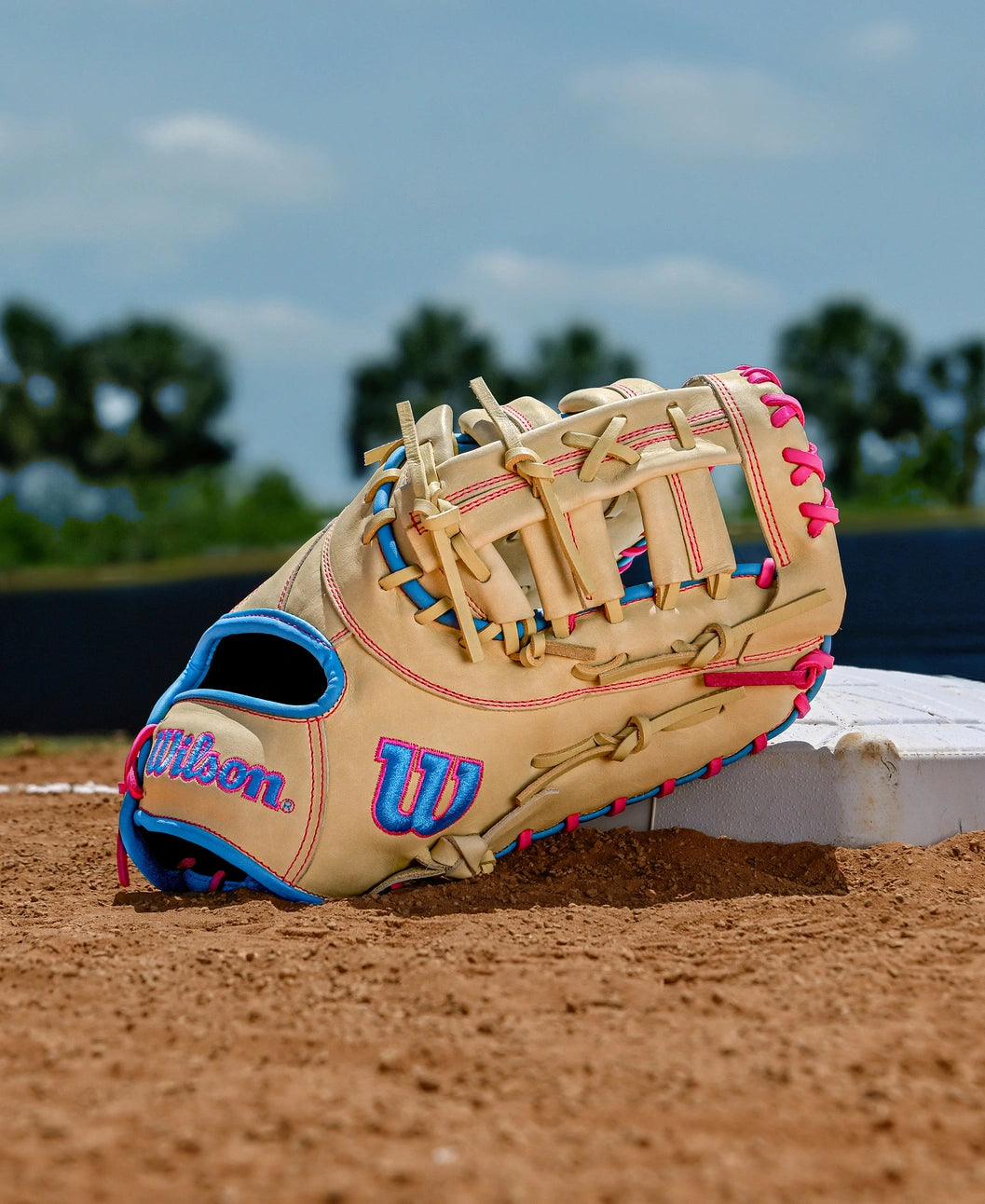 A Wilson A1000 1620 12.5" Baseball First Base Mitt by Wilson, in beige with blue and pink laces, sits on a base on a dirt field, framed by green trees and a blue sky in the background.