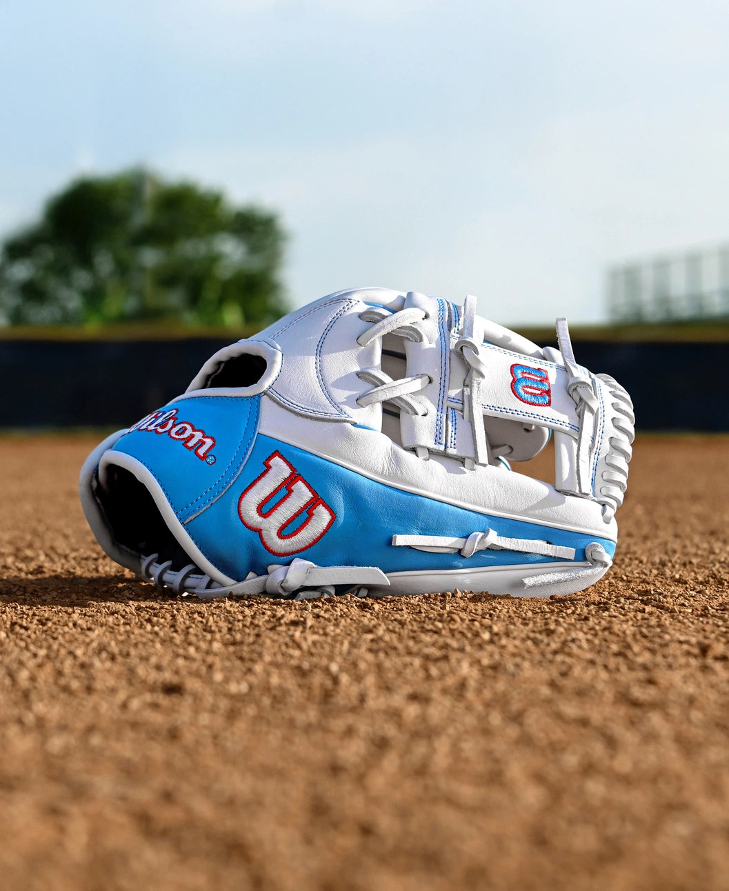 A Wilson A1000 H12 12" Fastpitch Glove (WBW10396912) by Wilson in white and blue rests on the infield dirt of a baseball field, with blurred trees and a fence visible in the background.