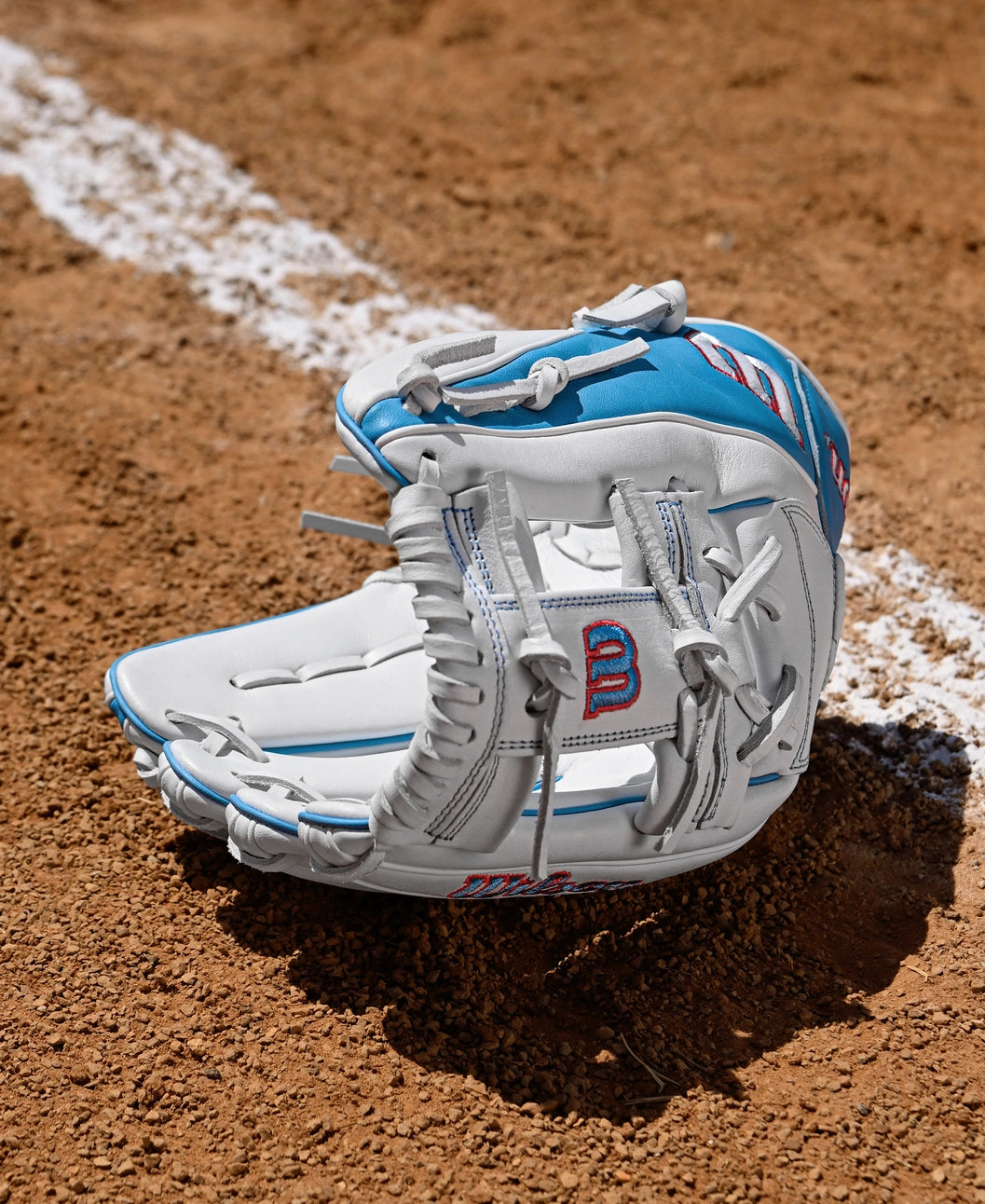A Wilson A1000 H12 12" Fastpitch Glove (WBW10396912) by Wilson in white and blue rests open on the dirt near a chalk baseline on the softball field.