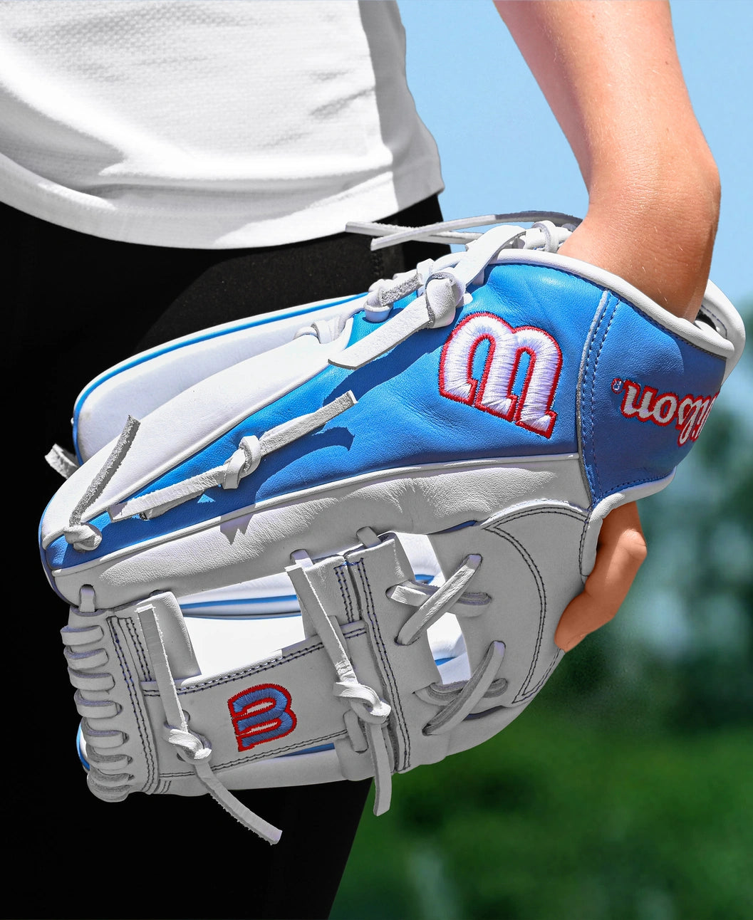 A person in black pants and a white shirt holds a Wilson A1000 H12 12" Fastpitch Glove (WBW10396912) outdoors, with a blurred green background.