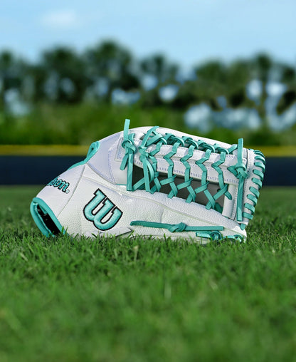 The Wilson A1000 T125 12.5" Fastpitch Glove by Wilson, in white and teal full grain leather, rests on green grass with a blurred background of trees and a dark fence.