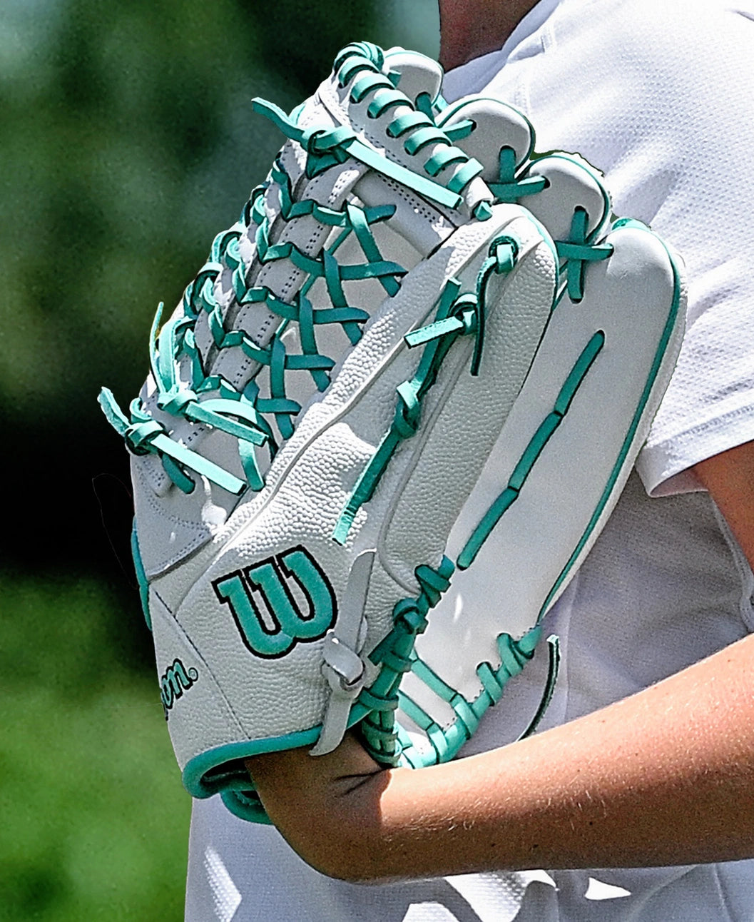 A person in a white shirt holds the Wilson A1000 T125 12.5" Fastpitch Glove (WBW103970125), a white youth glove with teal laces and the Wilson logo, made of full grain leather, with blurred green foliage in the background.