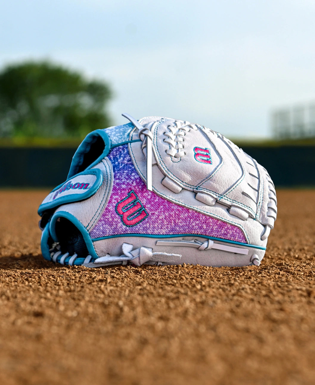 A Wilson Flash 11" Fastpitch Glove (WBW10398411), white and purple with teal accents, lies on a baseball field's dirt, green grass and a blurred tree in the background.