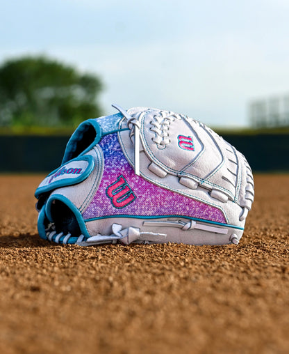 A Wilson Flash 11" Fastpitch Glove (WBW10398411), white and purple with teal accents, lies on a baseball field's dirt, green grass and a blurred tree in the background.