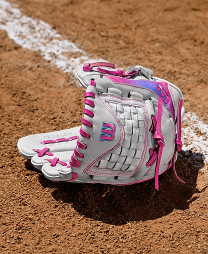 A Wilson Flash 11.5" Fastpitch Glove (WBW103985115) by Wilson in white and pink with blue stitching lies open on a dirt field beside a white chalk baseline.
