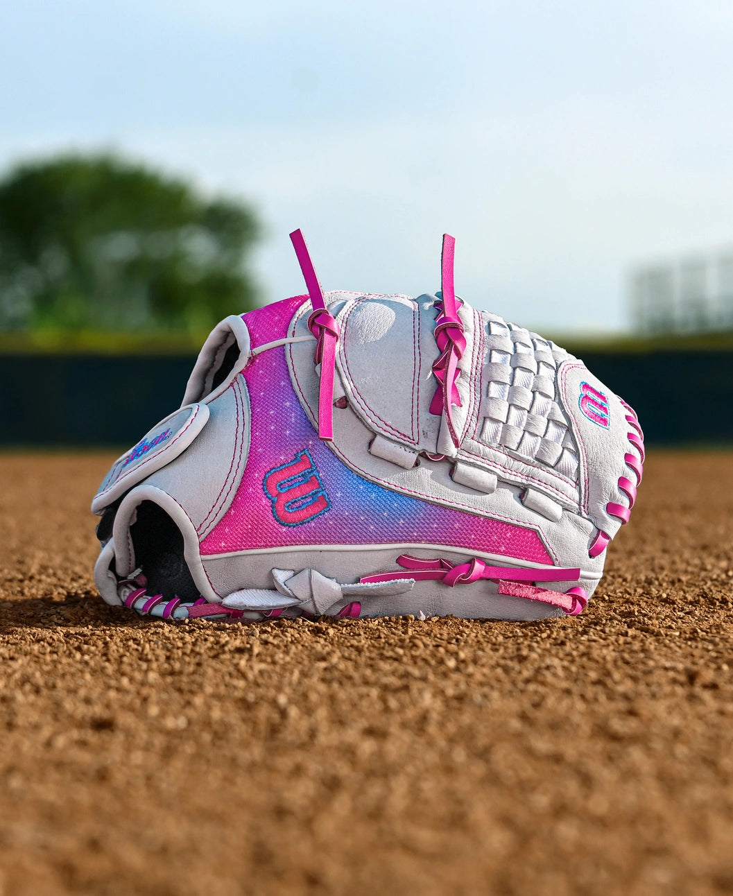 A Wilson Flash 11.5" Fastpitch Glove (WBW103985115), white and pink with the Wilson logo, rests on a dirt infield while green trees and an outfield fence blur in the background.