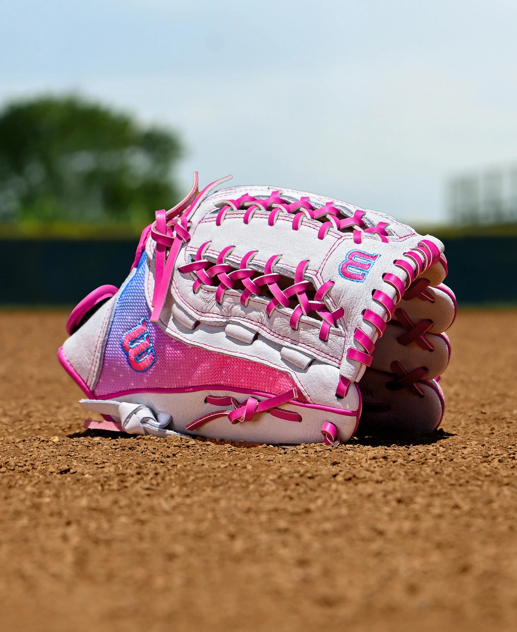 A Wilson Flash 12" Fastpitch Glove (WBW10398712) by Wilson rests on the dirt of a softball field; blurred trees and a fence can be seen in the background under a clear blue sky.