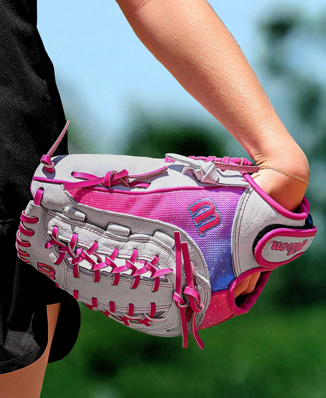 A person holds a Wilson Flash 12" Fastpitch Glove (WBW10398712) with gray, pink, and purple colors and bright pink laces outdoors on a sunny day. The glove is worn on their left hand.